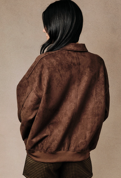 A person shown from the back wearing a Brown faux‑suede bomber jacket with modern silhouette and lightweight drape against a plain warm beige studio background.