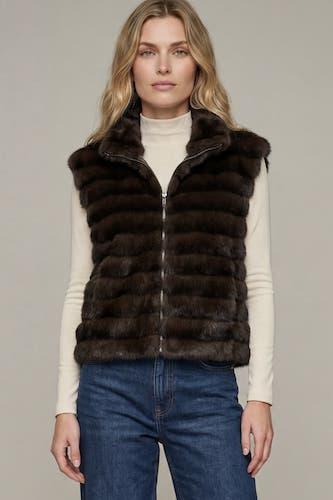 A woman wearing a dark brown fur-lined zip-front vest over a cream turtleneck and blue jeans, photographed against a plain light gray studio background.