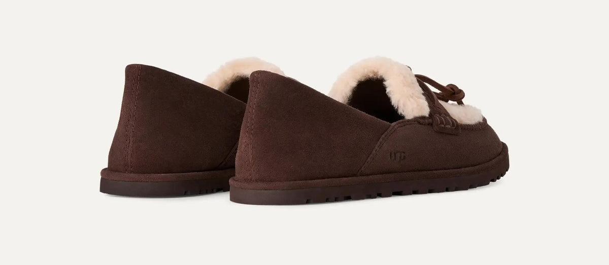 A pair of dark brown UGG Zora slip-on loafers with a soft cream shearling lining and a small bow detail on the front, shown against a plain off-white background.