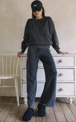 A woman models a dark gray washed crewneck sweatshirt and matching wide-leg pants, paired with a black Stussy cap and dark slippers, standing in a rustic room with a white wooden chest and chair.