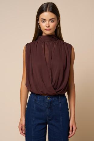 A woman modeling a sleeveless high-neck top in a deep plum-brown color, paired with dark blue jeans, against a solid beige background.