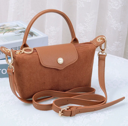 A brown suede handbag with a top handle, flap closure, and detachable shoulder strap, styled on a white textured surface with a decorative book in the background.