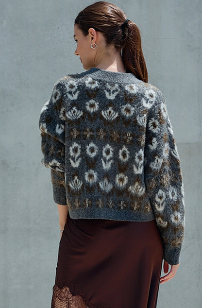 A woman shown from the back wearing a fuzzy knit cardigan with a gray, brown, and off-white floral pattern, paired with a dark brown skirt, set against a textured gray wall.