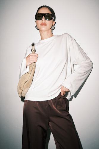 A model wearing a white long-sleeve top, dark brown wide-leg pants, black sunglasses, and a beige shoulder bag, standing against a light gray studio background.