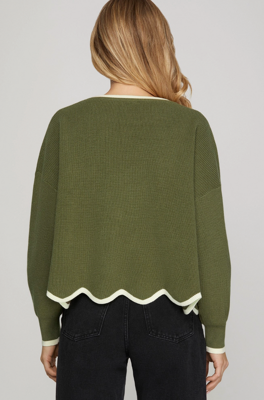 A woman viewed from the back wearing an olive green textured knit sweater with cream-colored ribbed trim at the crew neck, cuffs, and scalloped hem, paired with black jeans against a light gray studio background.