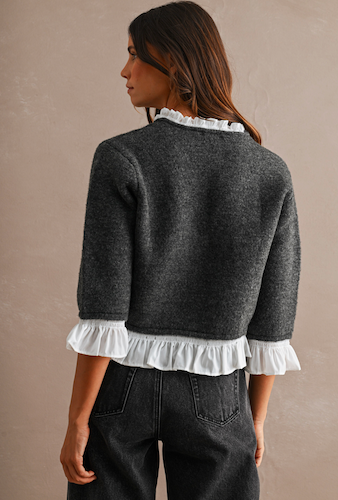 A woman shown from the back wearing a dark gray cropped sweater with white ruffled trim at the collar and cuffs, paired with dark jeans, against a plain beige studio background.