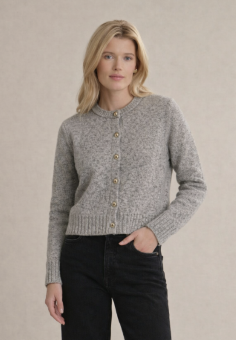 A woman modeling a light gray marled knit cardigan with gold buttons, paired with black jeans, against a neutral beige background.