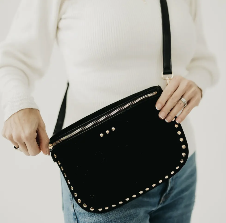 A person holding a black velvet crossbody bag with gold-tone studs and a zipper. The bag has a black strap and is shown against a plain white background.