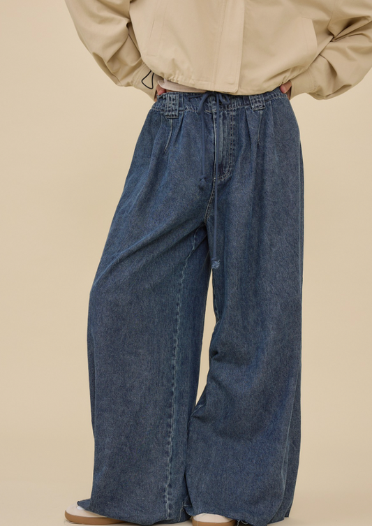 A person wearing loose, wide-leg blue denim jeans with a beige jacket and white shoes, shown from the shoulders down against a plain beige studio background.
