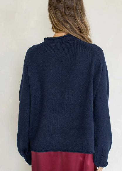 A woman viewed from the back wearing a dark navy blue knit sweater and a burgundy skirt, set against a plain off-white wall.