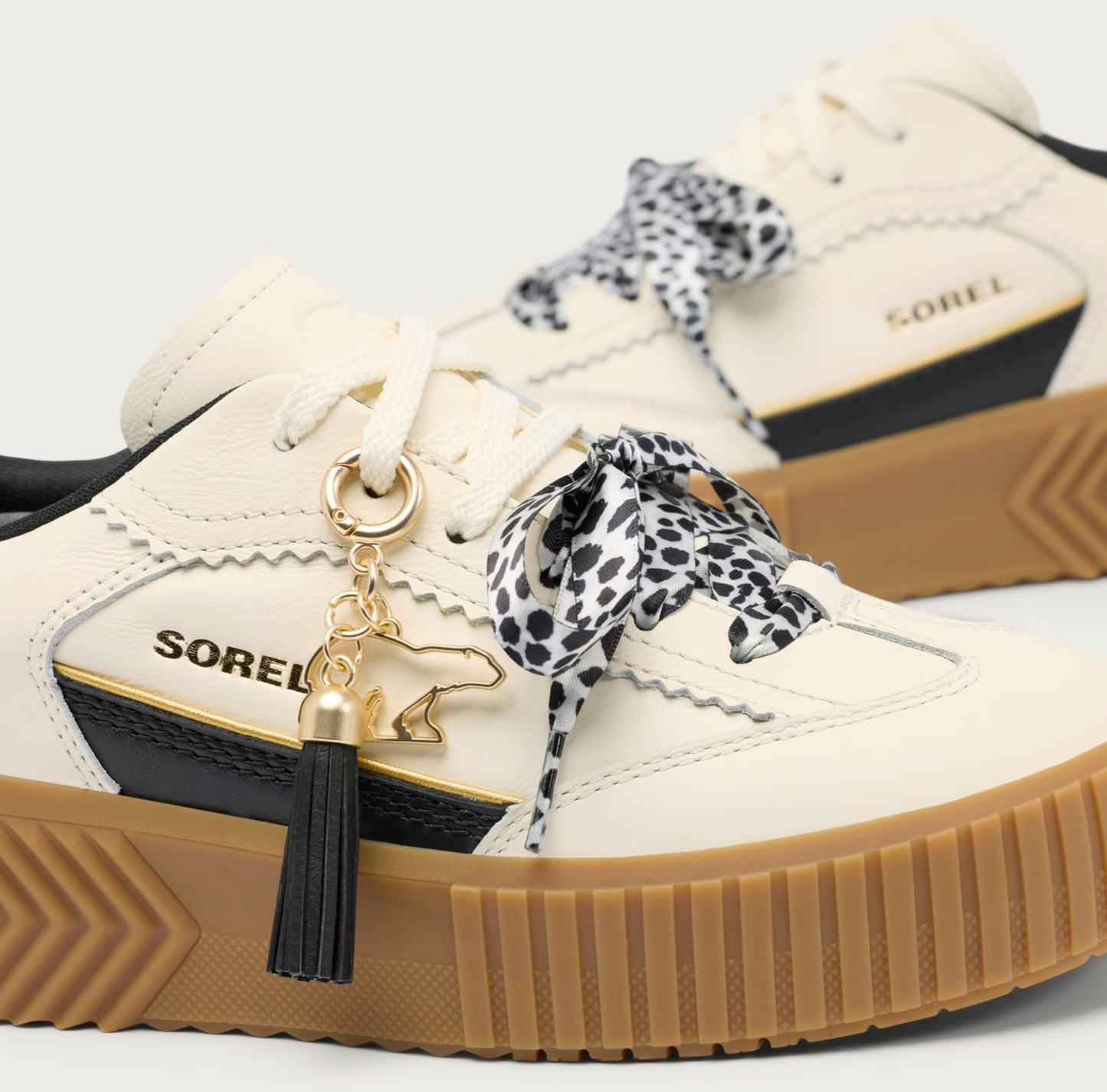 A pair of SOREL low-top sneakers in off-white leather with black and gold side panels, brown gum soles, and black-and-white leopard-print laces. A gold charm with a black tassel hangs from the lace of the foreground shoe.