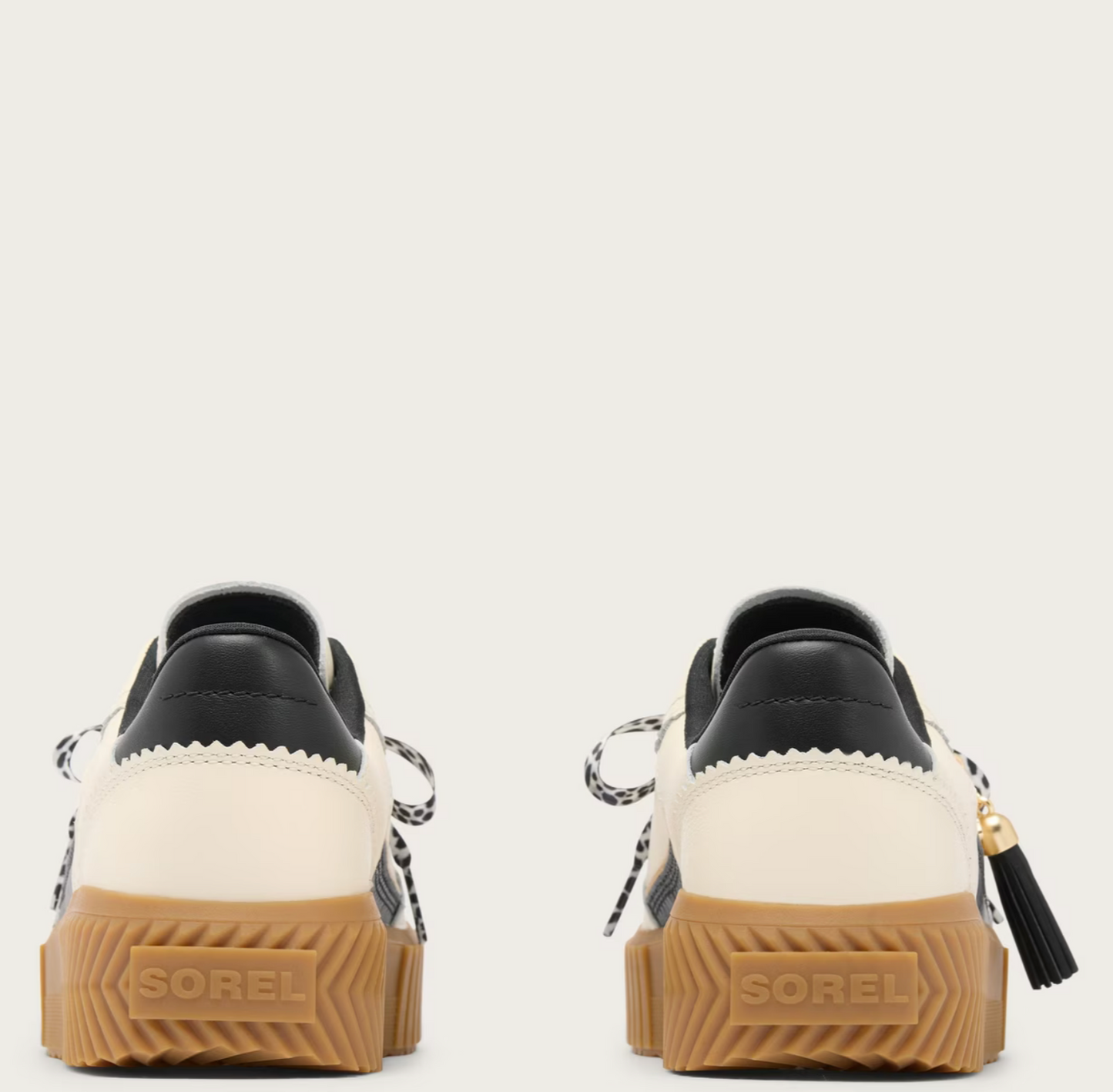 A pair of Sorel low-top sneakers shown from the back, featuring cream and black leather uppers, black-and-white patterned laces, and chunky brown gum soles with the Sorel brand name embossed.