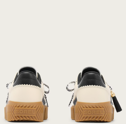 A pair of Sorel low-top sneakers shown from the back, featuring cream and black leather uppers, black-and-white patterned laces, and chunky brown gum soles with the Sorel brand name embossed.