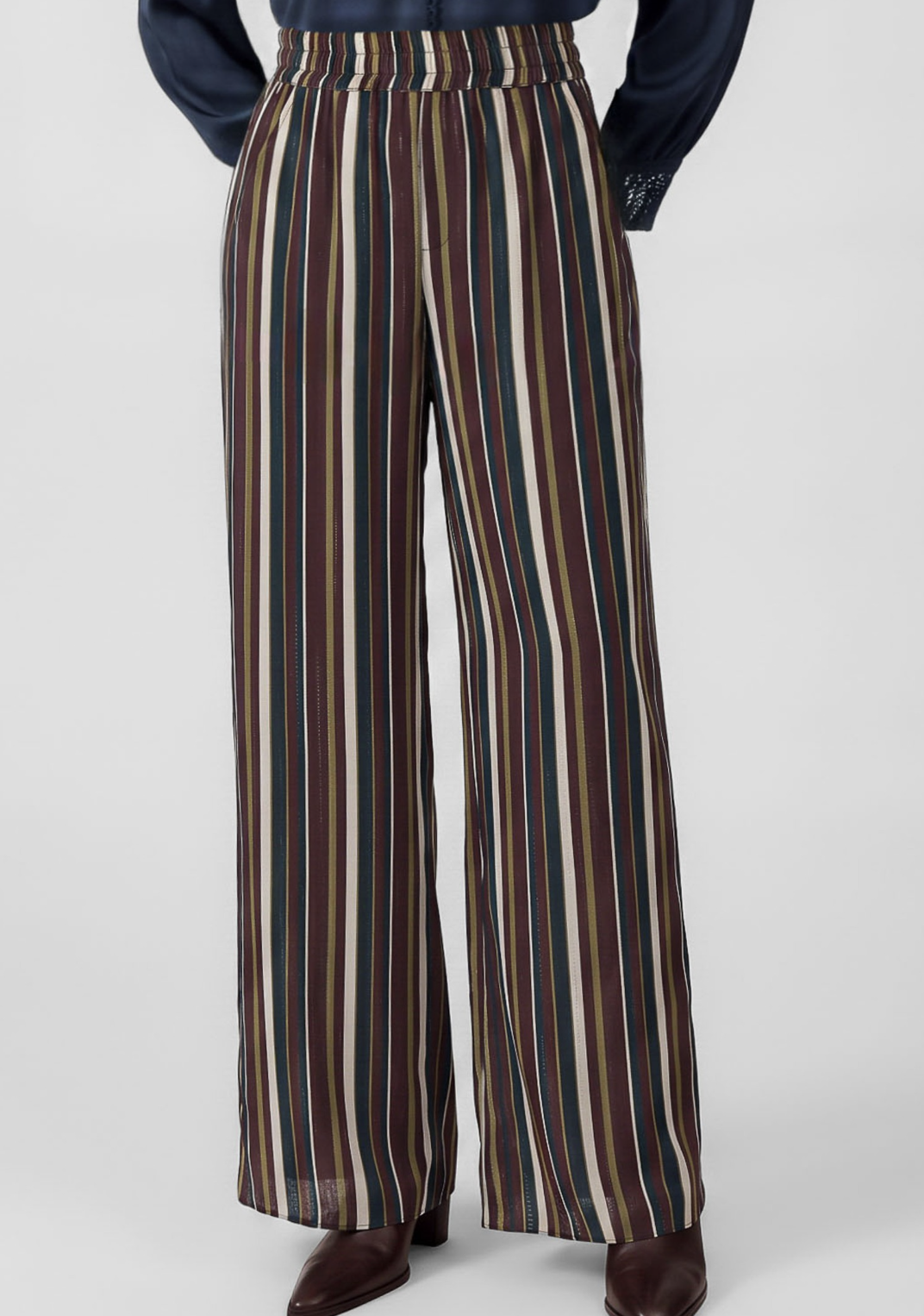 A person wearing high-waisted, wide-leg trousers with vertical stripes in brown, beige, cream, and dark green, paired with a navy top and brown shoes, shown from the waist down on a light gray studio background.