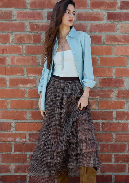 A woman stands against a red brick wall wearing a Brown plaid mesh maxi skirt with tiered ruffle layers and flowing silhouette..