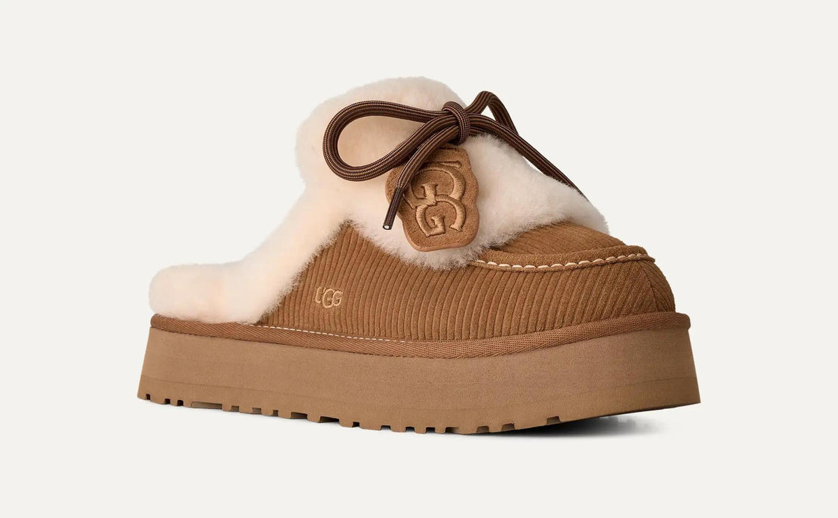 A UGG Disquette Heritage mule slipper in tan suede and brown corduroy with a thick platform sole, featuring a fluffy cream shearling lining, a brown bow, and a UGG logo patch on the side.
