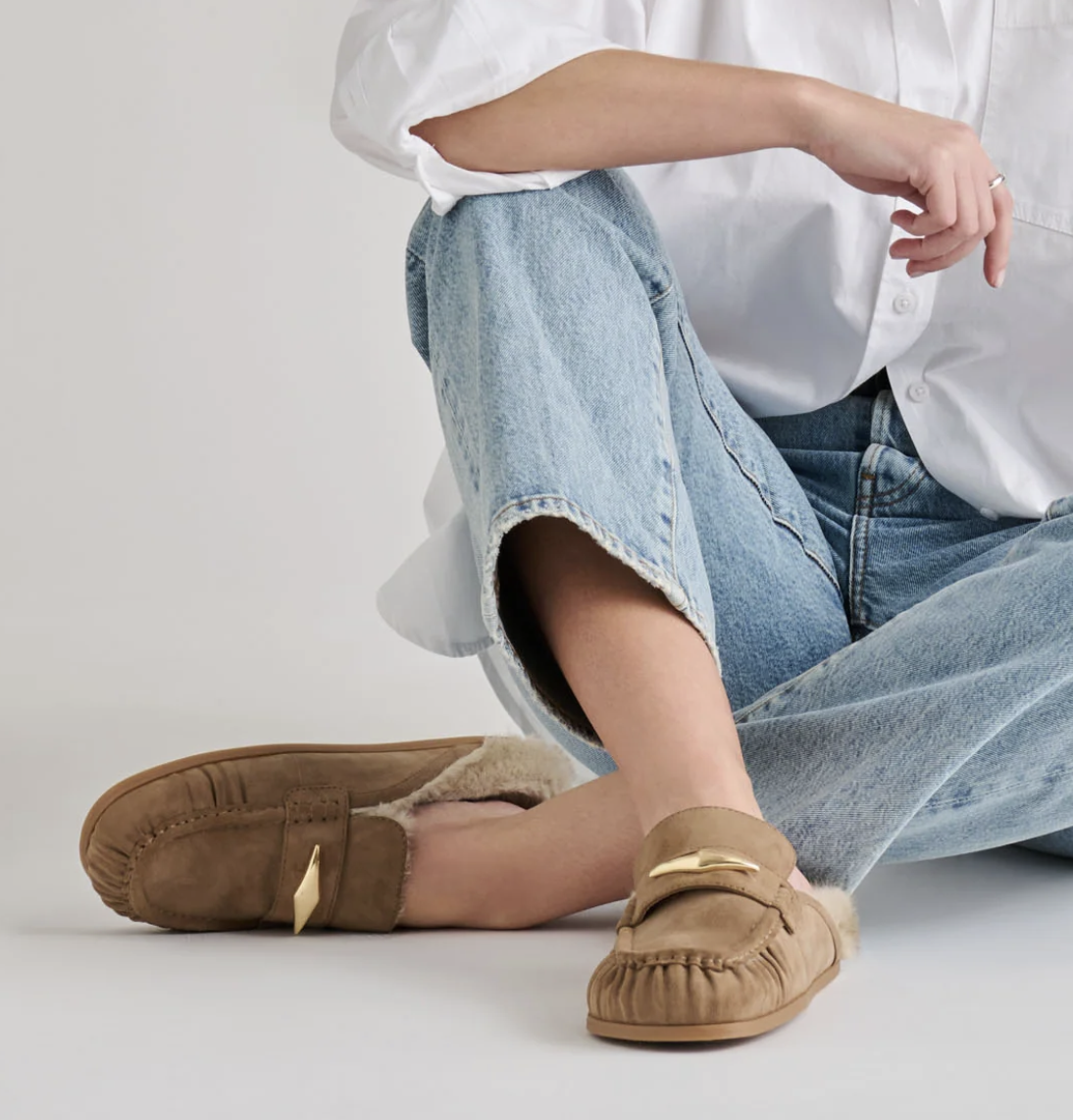 A person sitting on a light gray floor wearing Dolce Vita Jodero tan suede slip-on mule flats with a soft lining and a small gold-tone accent, paired with light blue jeans and a white button-up shirt.