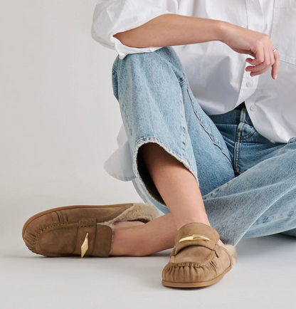 A person sitting on a light gray floor wearing Dolce Vita Jodero tan suede slip-on mule flats with a soft lining and a small gold-tone accent, paired with light blue jeans and a white button-up shirt.