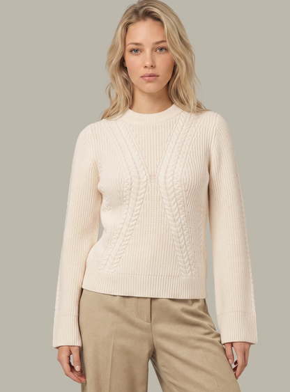A woman modeling a cream-colored 525 America cable-knit crewneck sweater with beige trousers, set against a solid light gray studio background.
