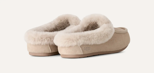 A pair of beige UGG Ansley slip-on mule with fluffy cream shearling lining, shown from a three-quarters angle on a light off-white background. The UGG logo is subtly embossed on the side of the heel.