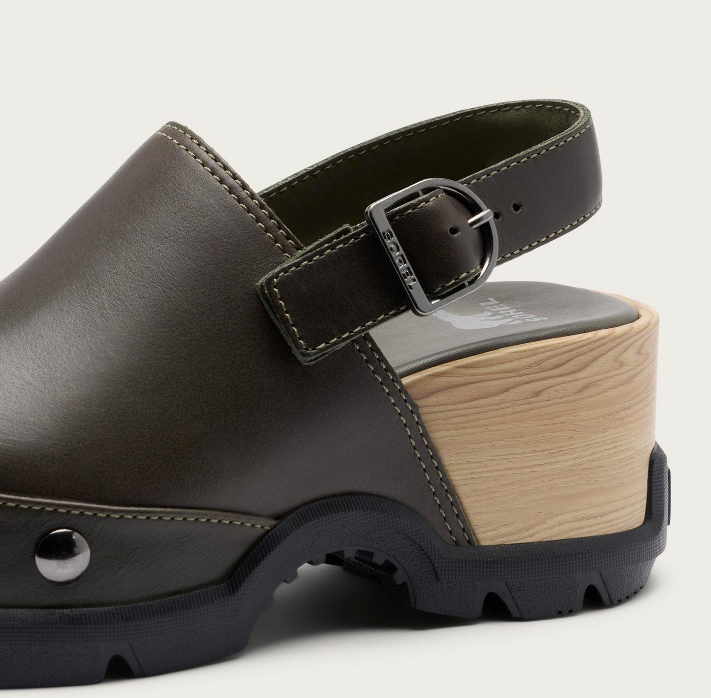 A close-up studio shot of a Sorel Roam’n clog-style mule shoe in dark brown leather with a light wood platform heel, black sole, and adjustable ankle strap with a silver buckle.