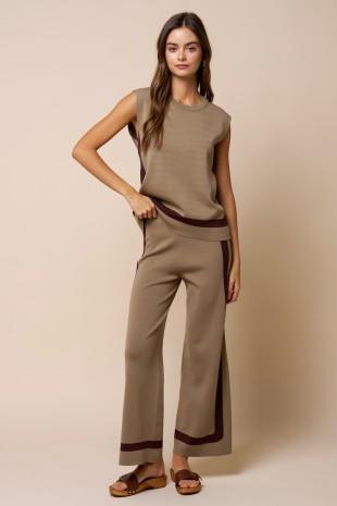 A woman modeling a sleeveless taupe knit top and matching wide-leg trousers with dark brown contrast trim, paired with brown slide sandals, against a solid beige studio background.
