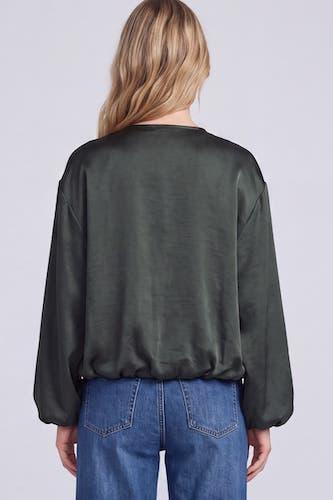 A woman viewed from the back wearing a dark olive green long-sleeve top and blue jeans against a light gray studio background.