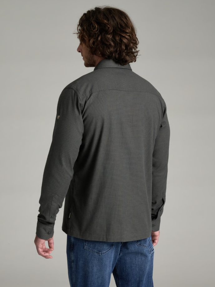 A man with long brown hair is shown from the back wearing a Kuhl Revolvr dark gray long-sleeve button-up shirt and blue jeans against a solid light gray studio background.