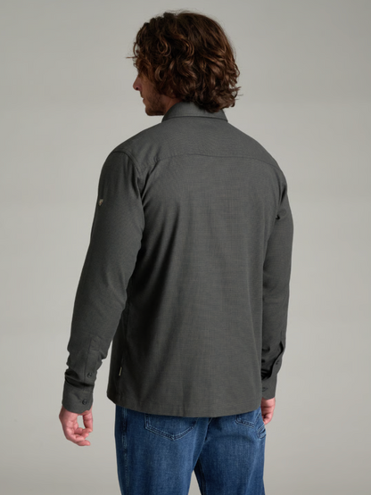 A man with long brown hair is shown from the back wearing a Kuhl Revolvr dark gray long-sleeve button-up shirt and blue jeans against a solid light gray studio background.