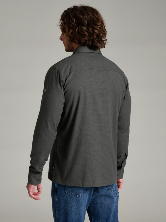 A man with long brown hair is shown from the back wearing a Kuhl Revolvr dark gray long-sleeve button-up shirt and blue jeans against a solid light gray studio background.