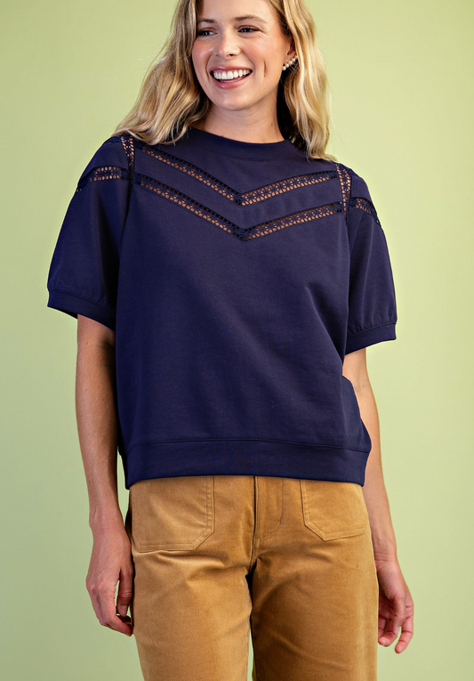 A woman wearing a navy blue short-sleeve crewneck sweatshirt with a decorative chevron lace detail across the chest, paired with tan pants, set against a solid light green background.