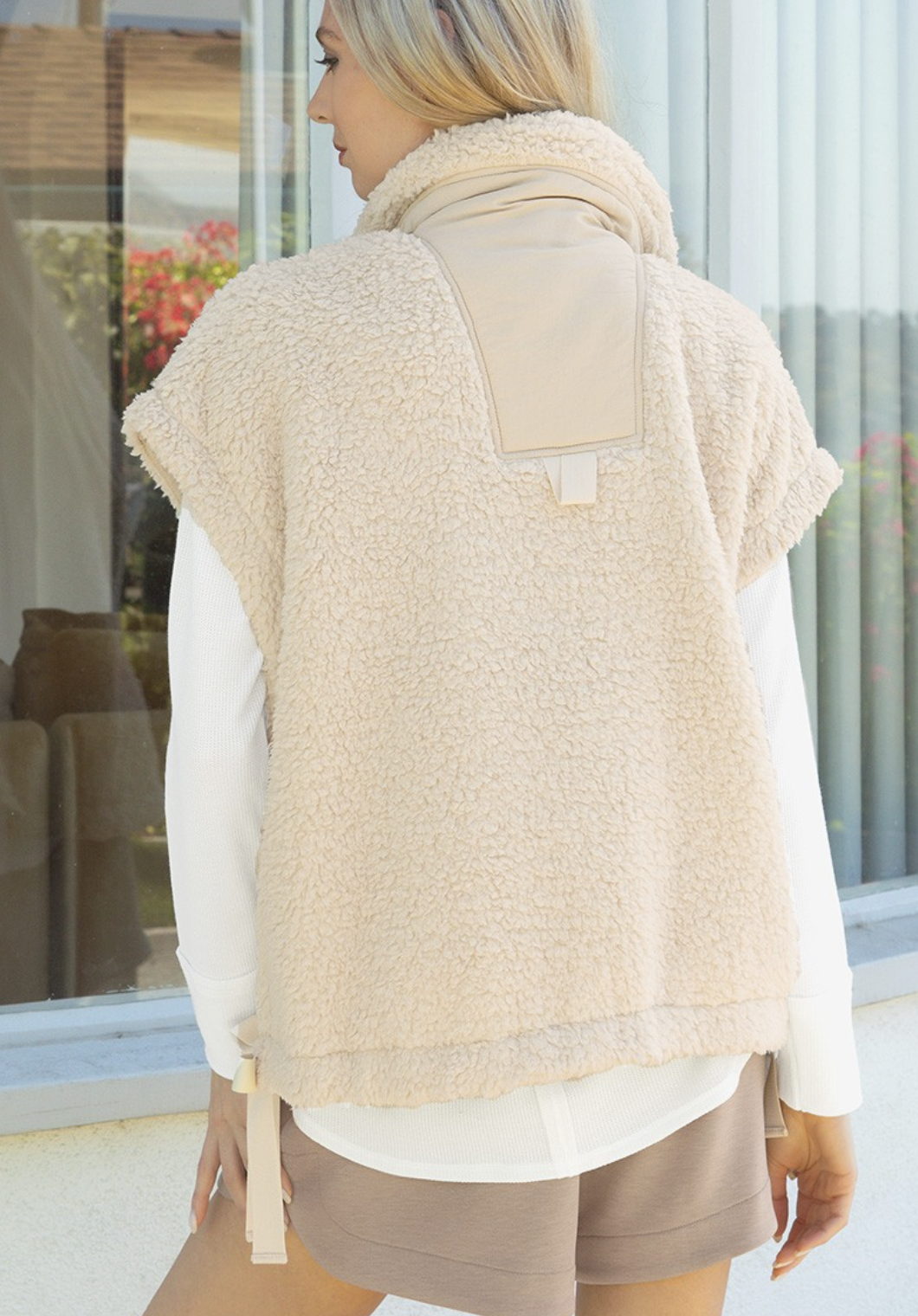 A woman shown from the back wearing a cream-colored sherpa fleece vest over a white long-sleeve shirt and taupe shorts, standing in front of a glass storefront window.