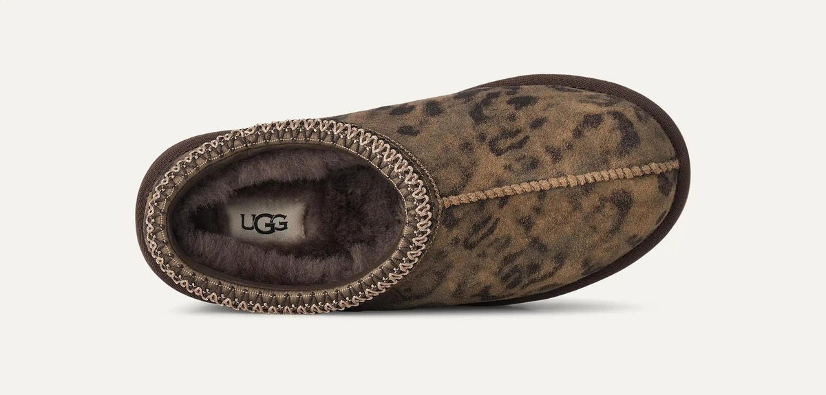 A single UGG Tasman Spotted slipper shown from a side profile view against a light off-white background. The slipper features a brown and black leopard print suede upper, a dark brown faux fur lining, and a decorative braided trim around the collar.