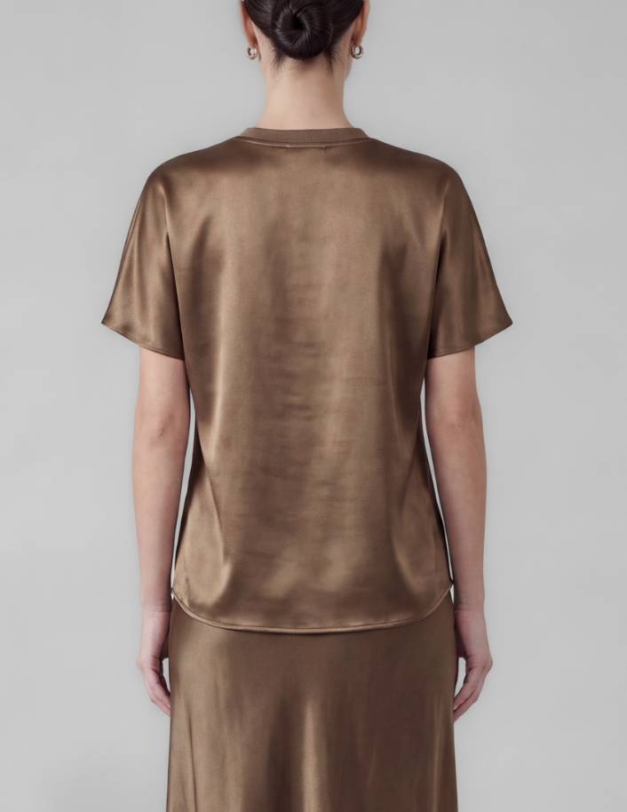 A woman viewed from the back wearing a short-sleeve satin-like top and matching skirt in a warm taupe-brown color against a plain light gray studio background.