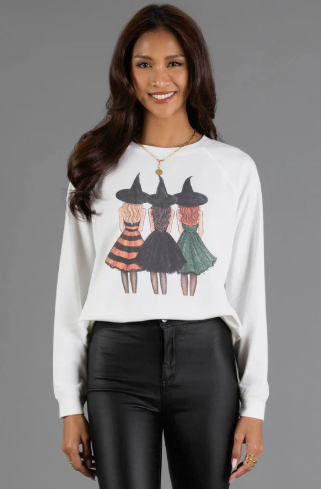 A woman modeling a white long-sleeve crewneck sweatshirt with a graphic of three witches wearing black pointed hats and colorful dresses, paired with black faux-leather pants, against a solid gray studio background.