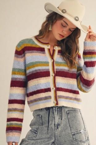 A woman wearing a LovLov cropped, long-sleeve knit cardigan with horizontal multicolor stripes in cream, yellow, green, blue, burgundy, and gray, paired with light-wash denim jeans and a cream cowboy hat.