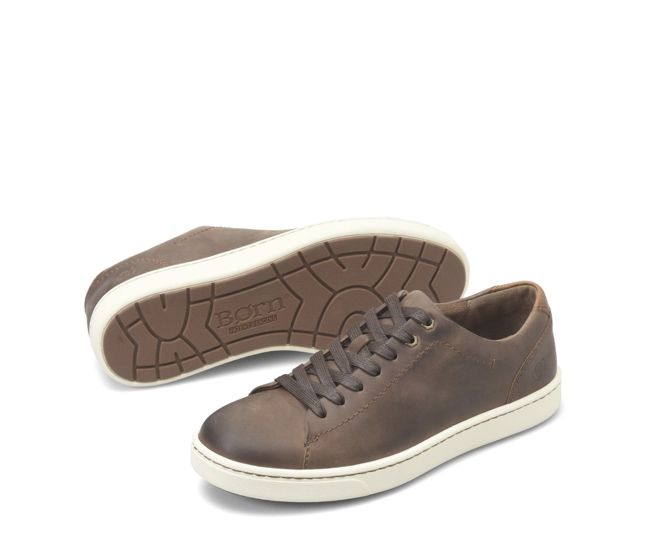 A pair of brown leather low-top sneakers, with one shoe standing upright and the other lying on its side to show the treaded outsole. The shoes feature a distressed brown finish, matching brown laces, and a thick cream-colored rubber sole.