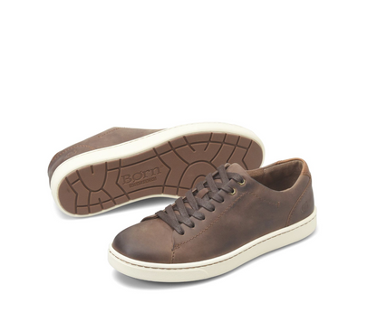 A pair of brown leather low-top sneakers, with one shoe standing upright and the other lying on its side to show the treaded outsole. The shoes feature a distressed brown finish, matching brown laces, and a thick cream-colored rubber sole.