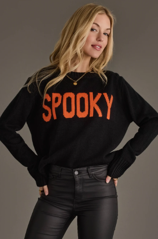 A woman modeling a black knit sweater with the word 'SPOOKY' in large orange letters across the chest, paired with black faux-leather pants, against a plain gray studio background.