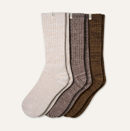Four UGG ribbed crew socks in neutral tones, fanned out on a light off-white background. The colors are light beige, heather gray, medium brown, and dark olive brown.