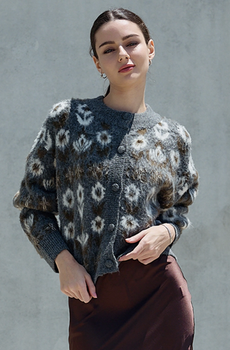 A woman wearing a fuzzy knit cardigan with a gray, brown, and white floral pattern, paired with a brown skirt, standing against a textured gray wall.