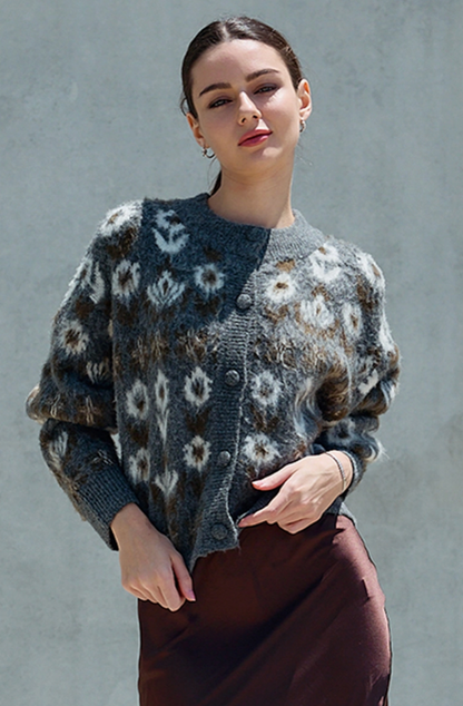 A woman wearing a fuzzy knit cardigan with a gray, brown, and white floral pattern, paired with a brown skirt, standing against a textured gray wall.