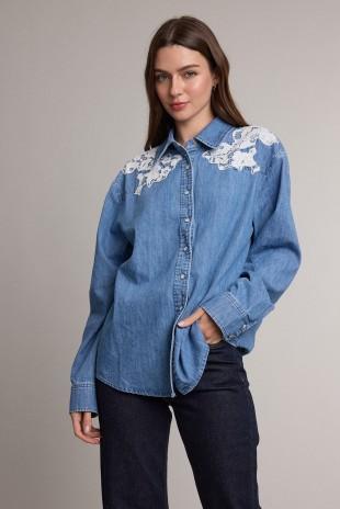A woman modeling a Raisonnel blue denim button-up shirt with white floral embroidery on the shoulders, paired with dark jeans, against a plain light gray studio background.