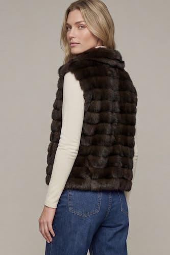 A woman shown from the back wearing a dark brown fur vest over a cream long-sleeve top and blue jeans, against a plain light gray studio background.