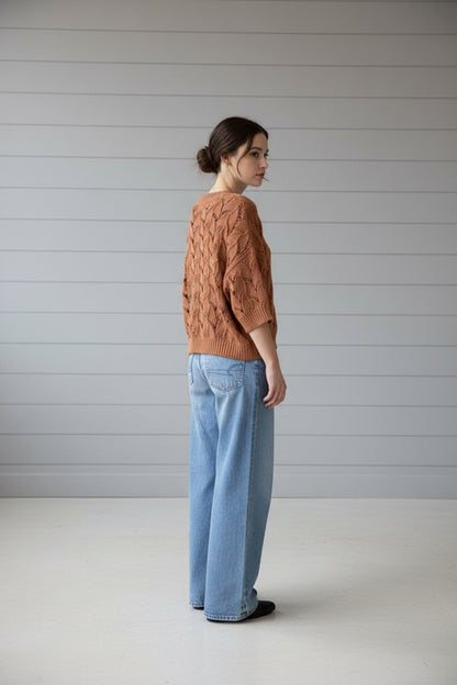 A person viewed from behind wearing a loose, textured knit sweater in a warm terracotta or rust color, paired with light blue jeans, standing outdoors next to a dark door and brick wall with metal mailboxes.