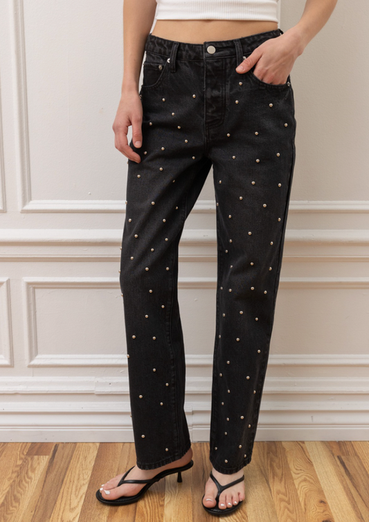 Black denim jeans with small white embroidered dots, modeled with a white crop top and black thong sandals against a white paneled wall and wood floor.