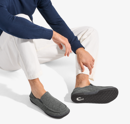 A man wearing a navy long-sleeve shirt and white pants sits on a white studio floor, showing a pair of Olukai Moloa Hulu gray felt slip-on loafers with black soles. One shoe is being adjusted with his hand.
