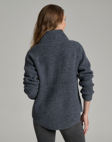 A woman viewed from the back wearing a dark heathered blue Kuhl Sienna ribbed knit sweater with a mock neck and long sleeves, paired with dark gray pants, against a solid gray studio background.