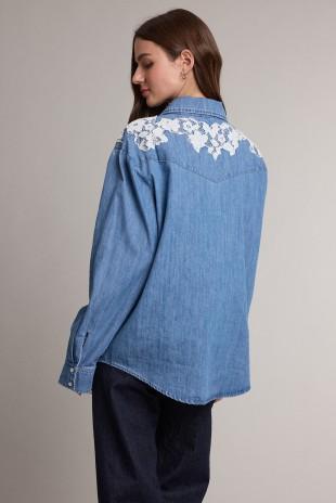A woman shown from the back wearing a light blue Raisonnel denim shirt jacket with white floral lace embroidery across the shoulders, paired with dark pants, against a plain light gray studio background.