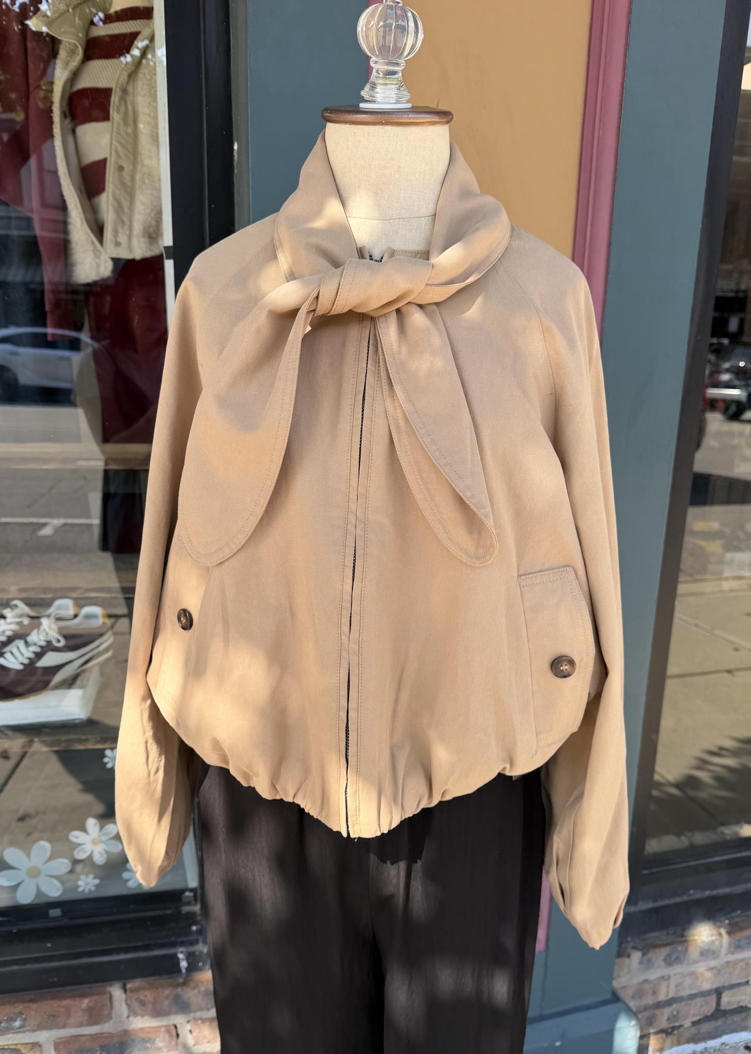 A beige cropped Entro jacket with a large knotted scarf detail at the collar, displayed on a mannequin torso with black pants in front of a storefront window.